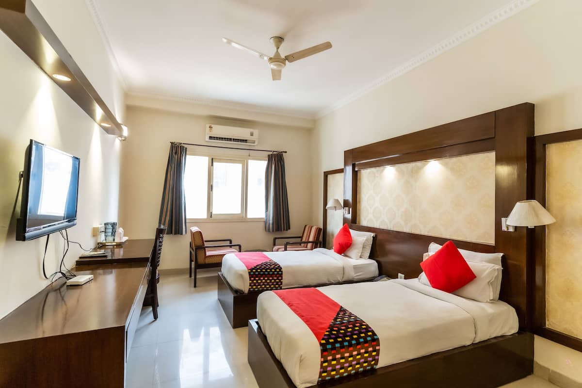 A hotel room with two beds, a TV, a desk, a chair, a ceiling fan, and a window with curtains.