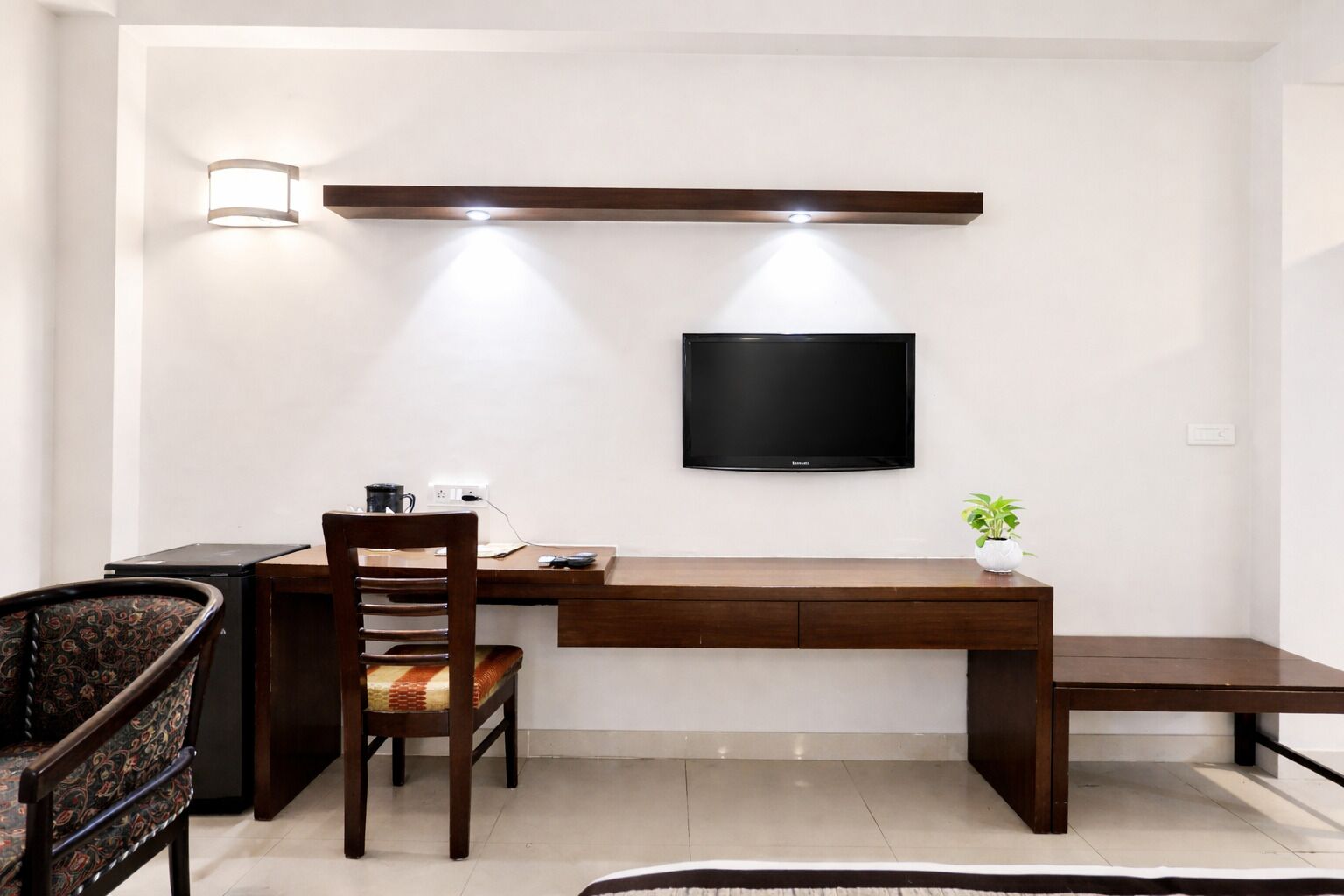 Een moderne kamer met een houten bureau, een stoel, een aan de muur bevestigde televisie en een kleine plant in pot.