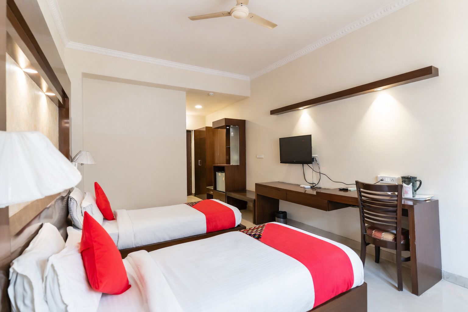 A hotel room with two beds, a desk with a chair, a TV, and a ceiling fan.