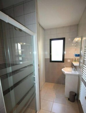 Ein modernes Badezimmer mit Duschkabine aus Glas, Waschbecken mit Spiegel und einer kleinen Mülltonne.