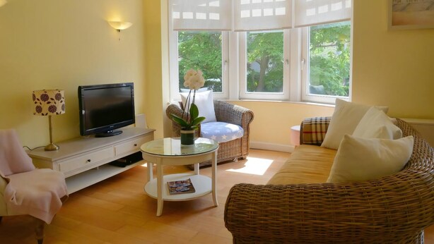 A living room with a television, a glass table, wicker furniture, and large windows with a view of trees.
