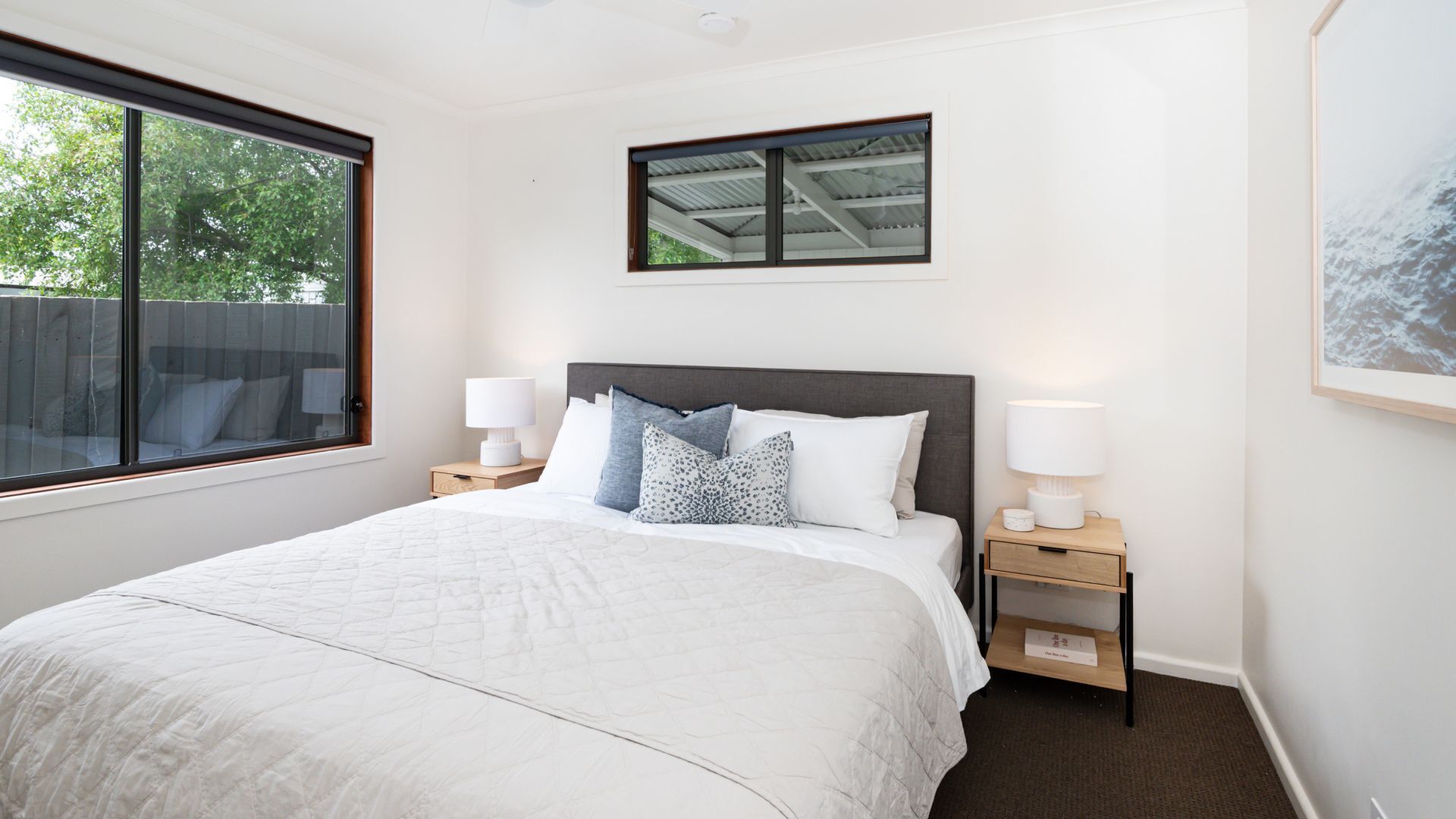 A bedroom with a large bed, two bedside tables with lamps, a window with a view of trees, and a framed picture on the wall.