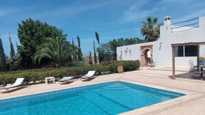 A swimming pool with lounge chairs, surrounded by greenery and a white building with an archway.
