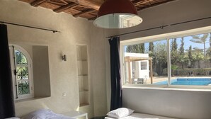 A bedroom with two beds, a large window with a view of a swimming pool and outdoor seating, and a ceiling with exposed wooden beams.