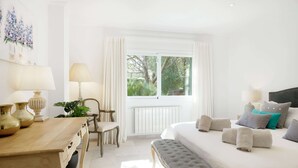 Een lichte, goed verlichte slaapkamer met een groot bed, nachtlampjes, een houten tafeltje met lampen en uitzicht op groen buiten het raam.
