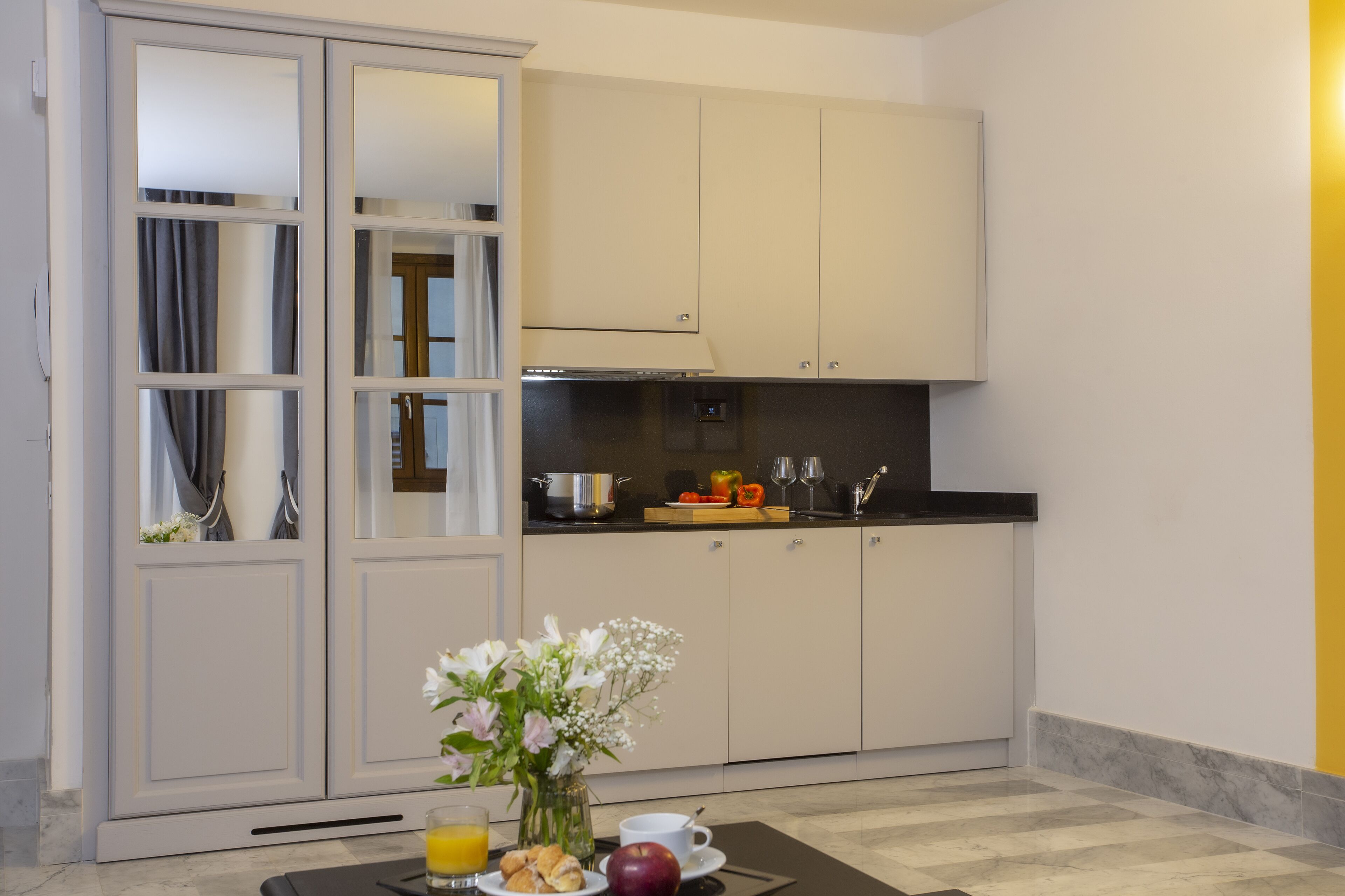 A modern kitchen with white cabinets, a black countertop, and a breakfast setup including croissants, a red apple, and a vase of flowers.