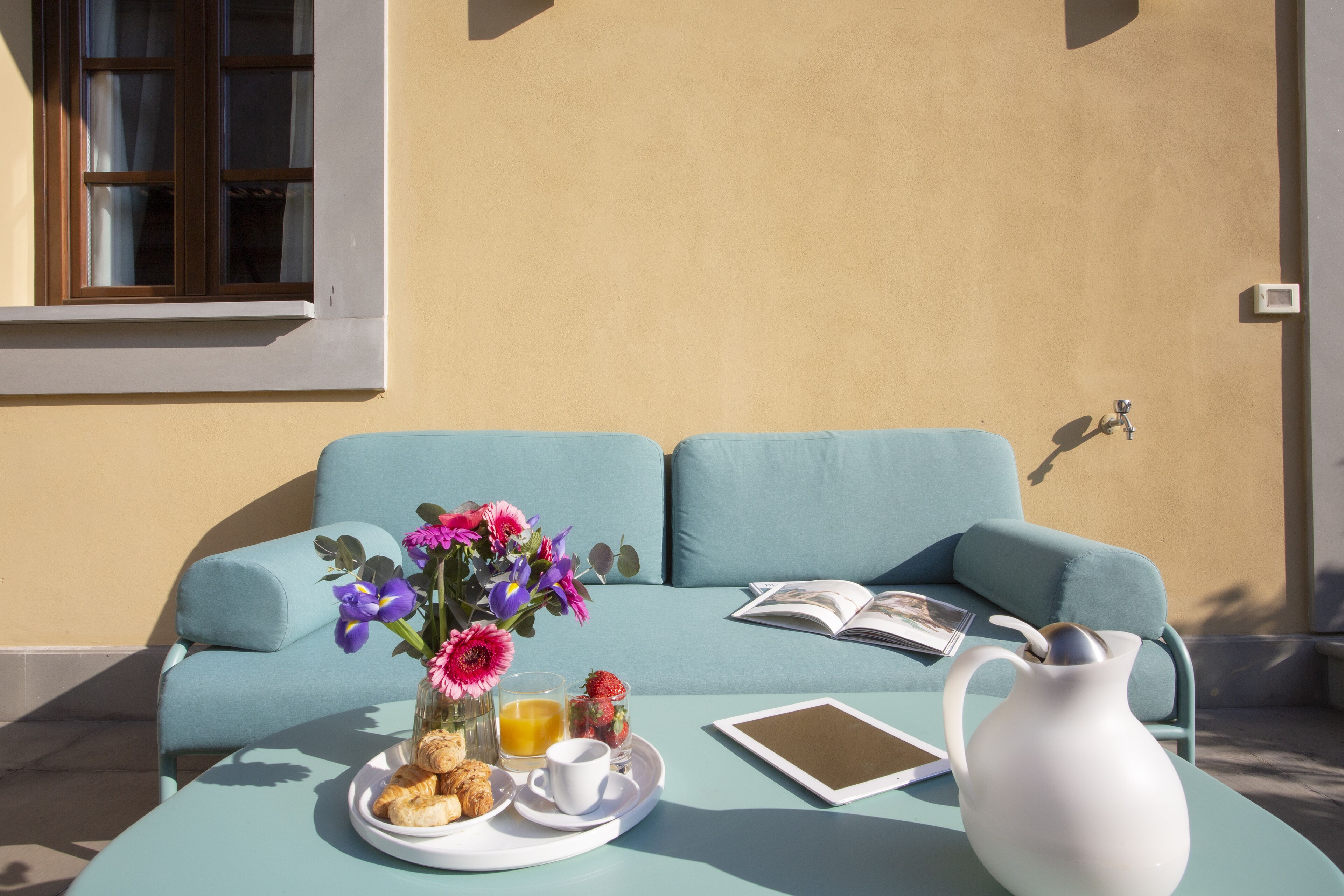 A teal sofa with a book and a tablet, a white pitcher, a plate with croissants, a cup, and a glass of orange juice on a round table.