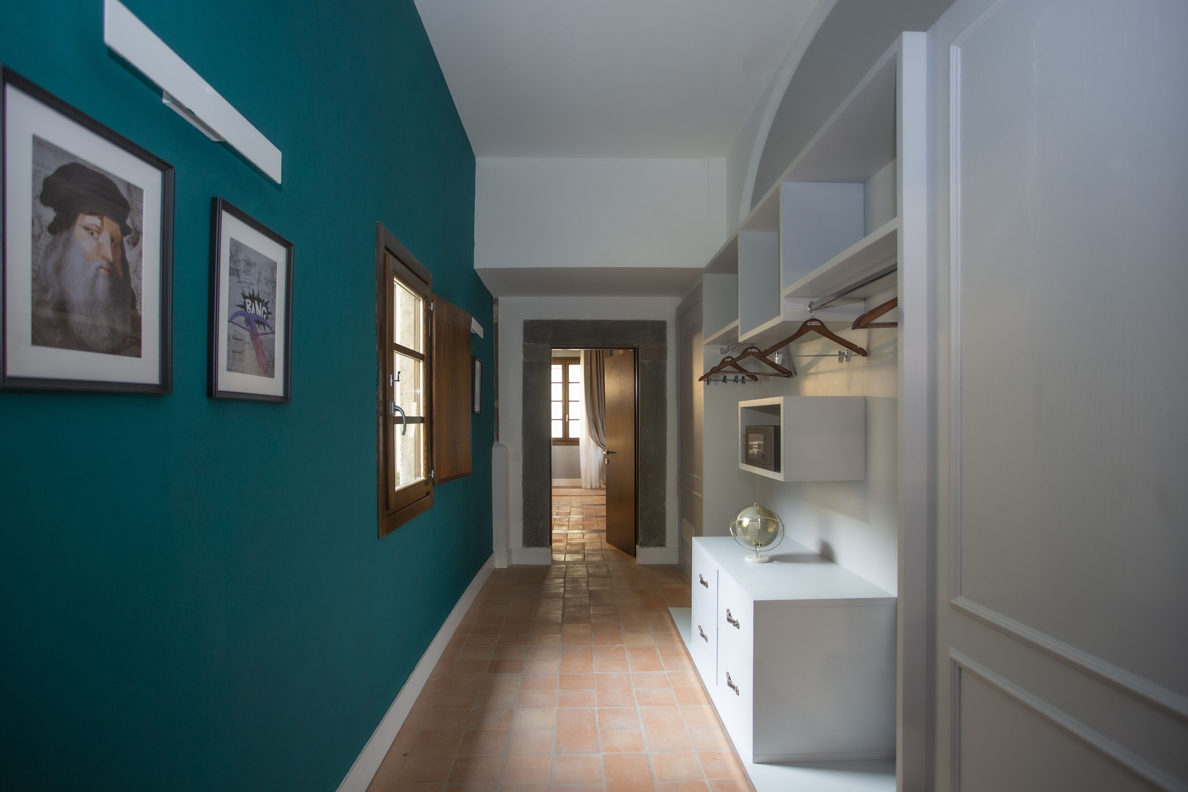 Un couloir aux murs de couleur turquoise, avec des portes en bois et un sol carrelé. Des tableaux encadrés ornent les murs, et une console blanche supporte un globe terrestre.