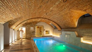 Une piscine intérieure avec un plafond en briques et des murs en pierre, une personne qui passe, et une porte vitrée donnant sur une autre pièce.