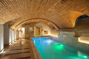An indoor swimming pool with a brick ceiling and stone walls, a person walking by, and a glass door leading to another room.