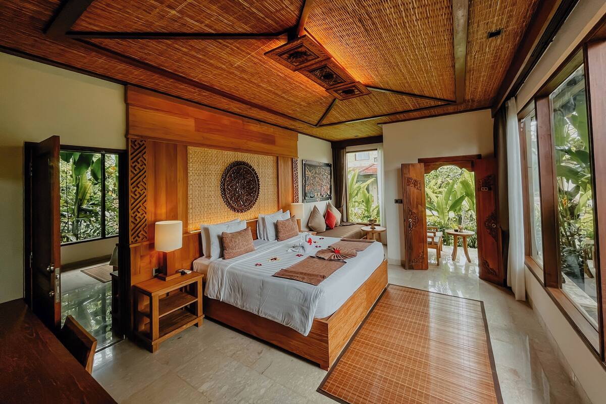 A spacious bedroom with a large bed, wooden headboard, and a view of lush greenery through the open door.
