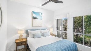 Um quarto com uma cama, duas mesas de cabeceira com luminárias, um ventilador de teto, uma janela com vista para árvores e um quadro na parede.