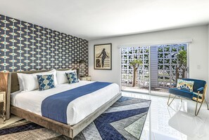 Een slaapkamer met behang met een patroon, een bed met meerdere kussens, een nachtkastje, een lamp, een ingelijst schilderij aan de muur en uitzicht door een schuifdeur naar een buitenruimte met een palmboom.