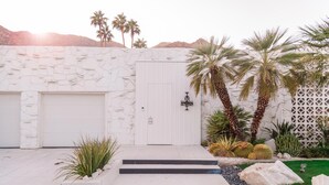 Une maison moderne avec une façade en pierre, deux portes de garage, un petit jardin agrémenté de cactus et de palmiers, et une vue dégagée sur les montagnes en arrière-plan.
