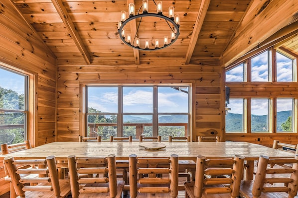 Enjoy meals in a spacious dining area with a large round table and comfortable chairs, surrounded by a high ceiling and large windows offering views of the surrounding landscape.