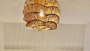 Un lit bien fait, avec des draps blancs, une couverture à carreaux aux couleurs vives et un luminaire original en fibres tressées suspendu au-dessus.