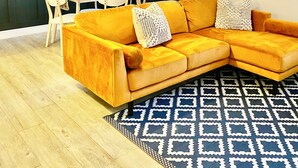 Un salon avec un canapé jaune, une table à manger avec des chaises, une cuisine visible à travers une fenêtre et des tableaux encadrés accrochés au mur.