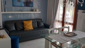 A modern living room with a dark grey sofa, a yellow pillow, a glass table with a metal tray, and a white side table with a speaker and a small item on it.