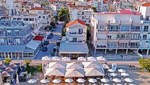 A coastal town with a beach area featuring sun loungers and umbrellas, a street with parked cars, and multi-story buildings with balconies. - Eantos Studios and Suites (Thermaikos)