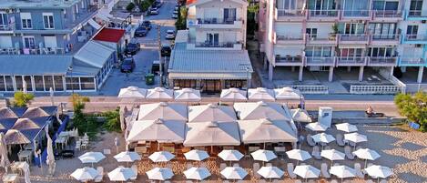 Een kustplaats met een strandgebied, ligbedden en parasols, een straat met geparkeerde auto's en meerhoogbouw met balkons.