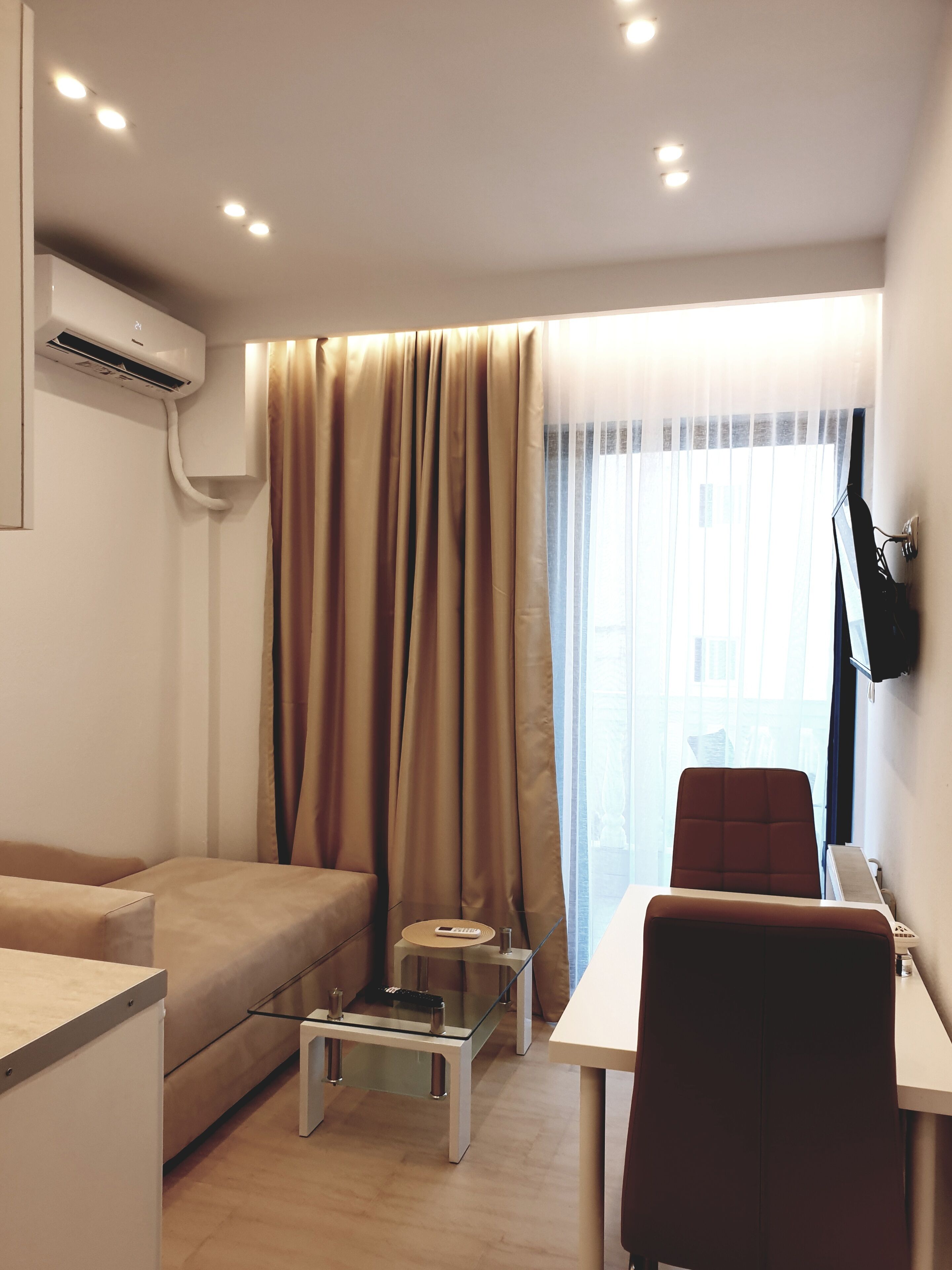 A modern living room with a sofa, a glass coffee table, a red chair, a TV mounted on the wall, and a large window with sheer curtains.