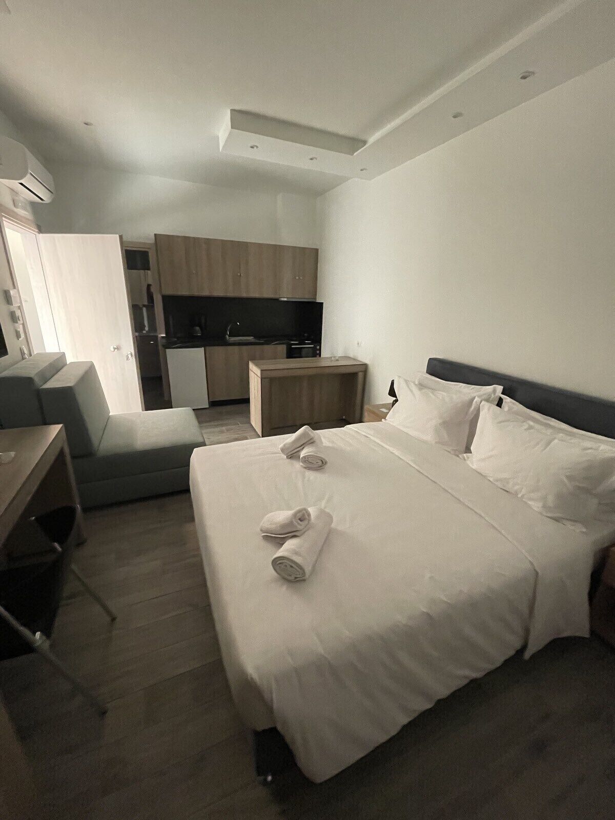 A modern hotel room with a neatly made bed, a small desk, and a kitchenette in the background.