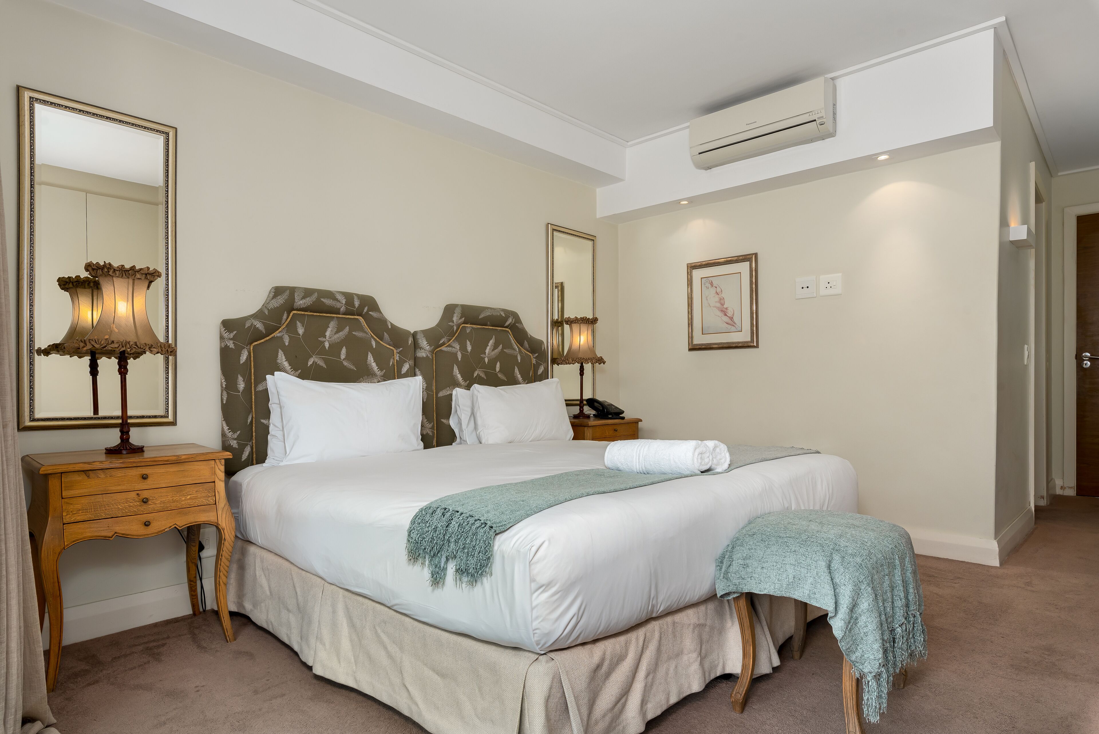 A neatly made bed with a green throw, two bedside lamps, a mirror, and a nightstand.