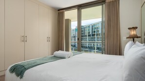 A hotel room with a large bed, a green throw blanket, a window with curtains, and a view of buildings outside. - Canal Quays 607 (Cape Town)