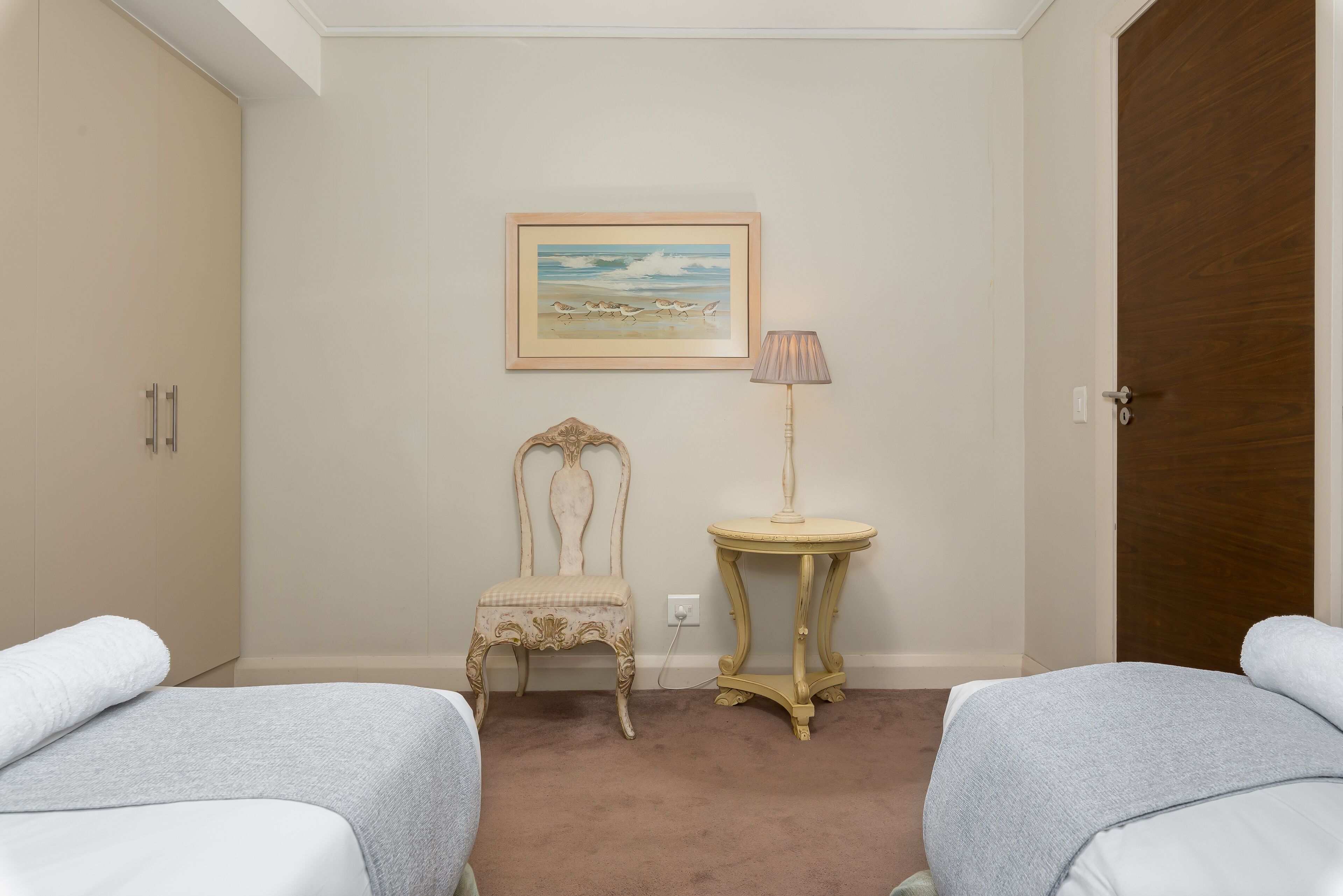 Habitación de hotel con dos camas, una puerta de madera, un cuadro enmarco, una lámpara y una silla decorativa.