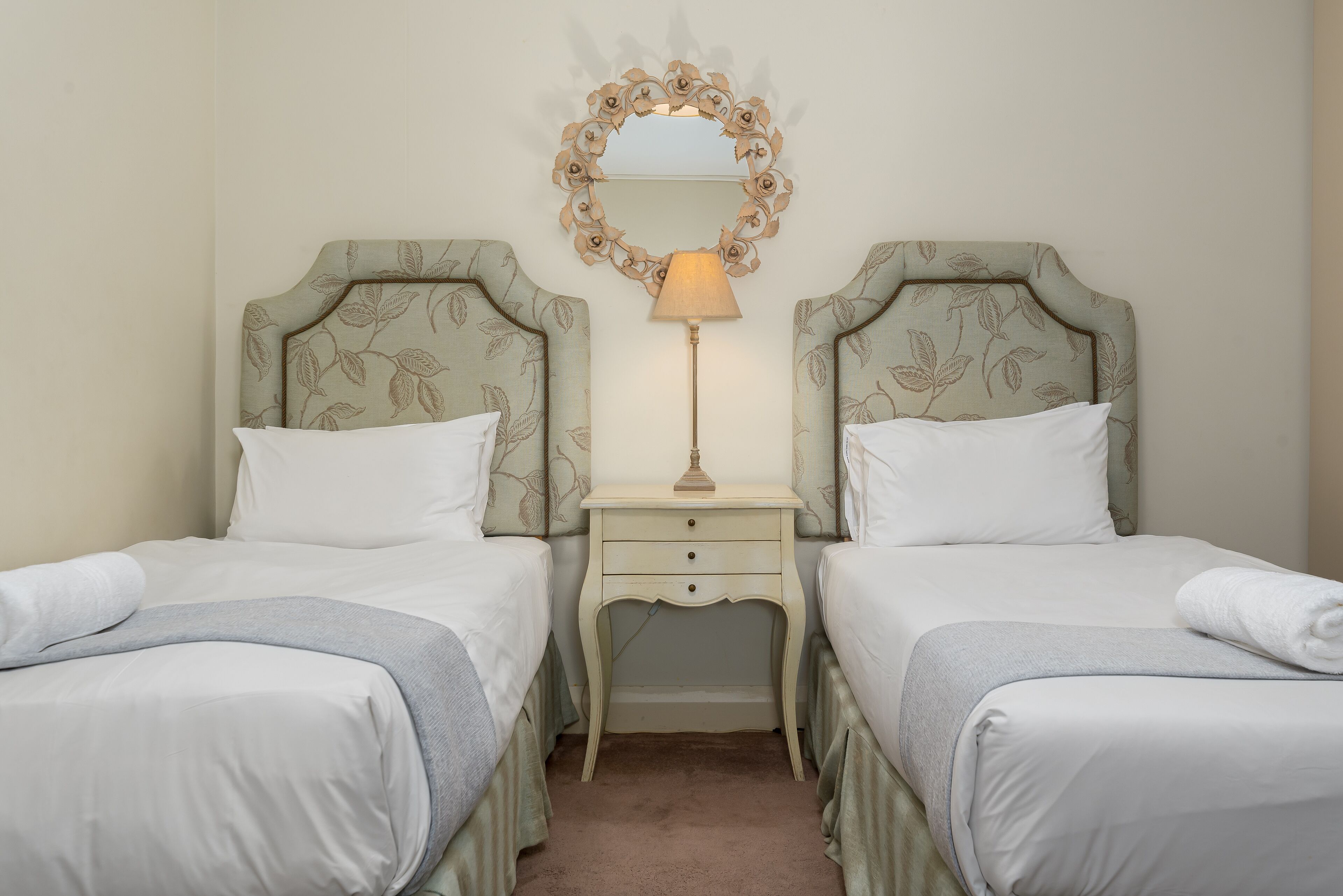 A hotel room with two beds, a nightstand with a lamp, and a decorative mirror above the beds.