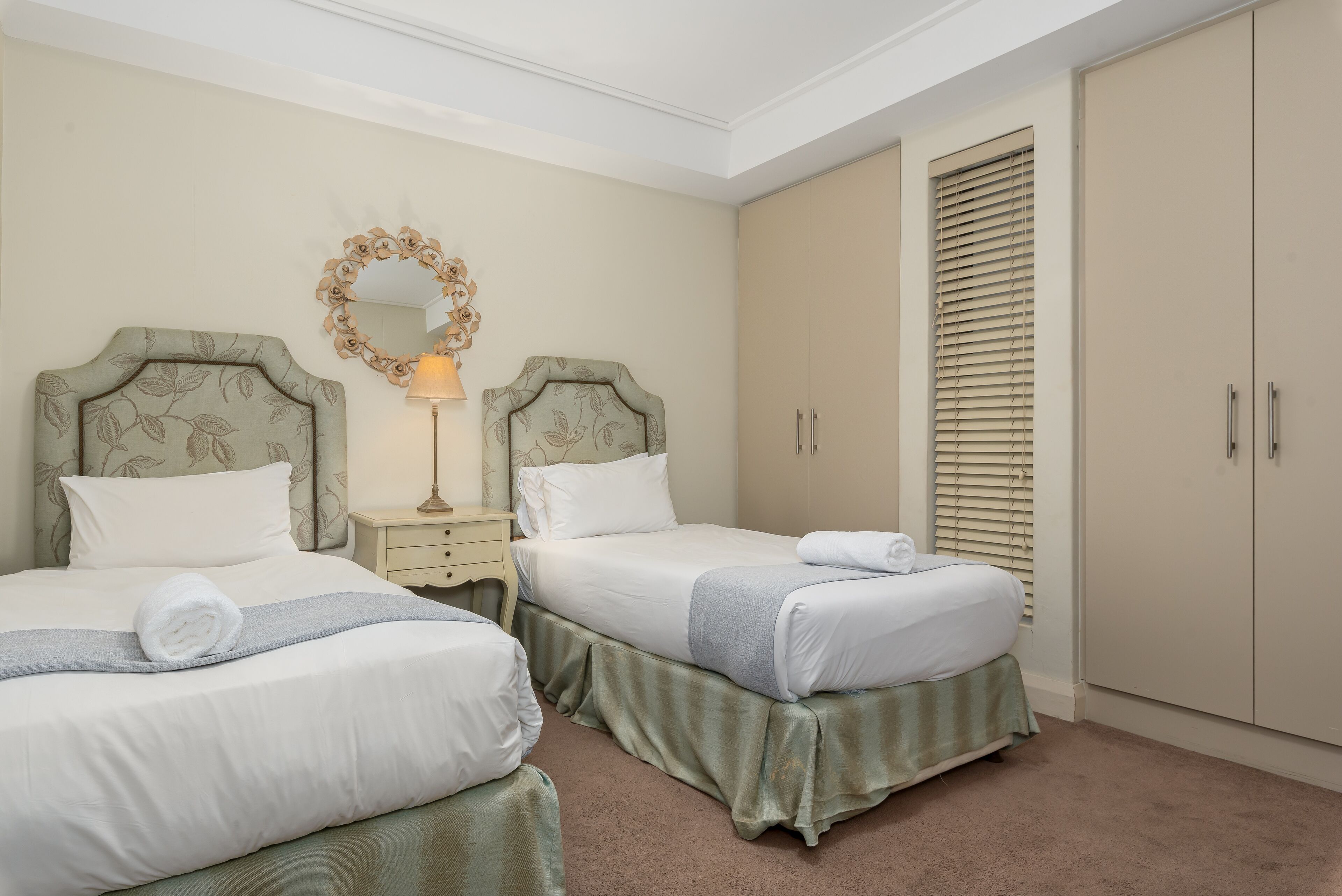 A hotel room with two beds, a nightstand with a lamp, a mirror, and a wardrobe.