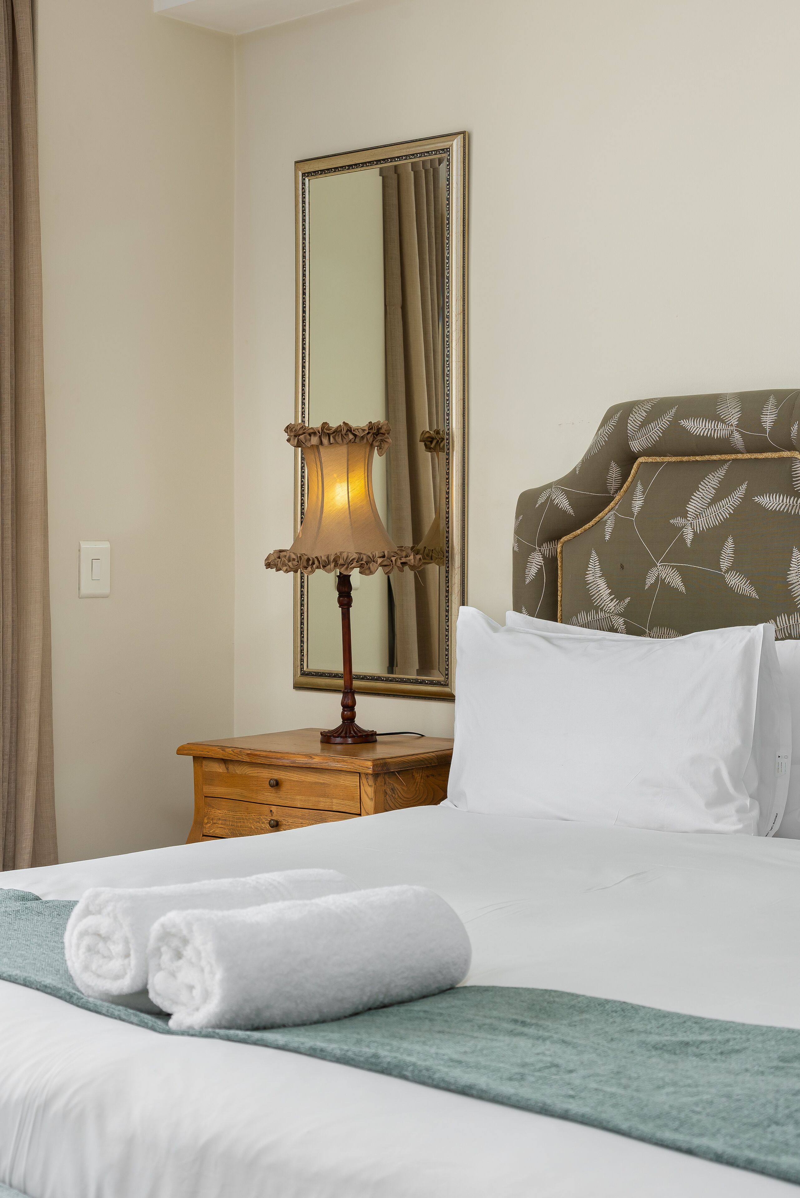 A neatly made bed with white linens, a green throw blanket, and two rolled white towels. There is a wooden nightstand with a lamp and a mirror.