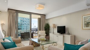 A modern living room with a glass-top table, beige sofas, teal cushions, a television, and a view of a building through large windows. - Canal Quays 607 (Cape Town)