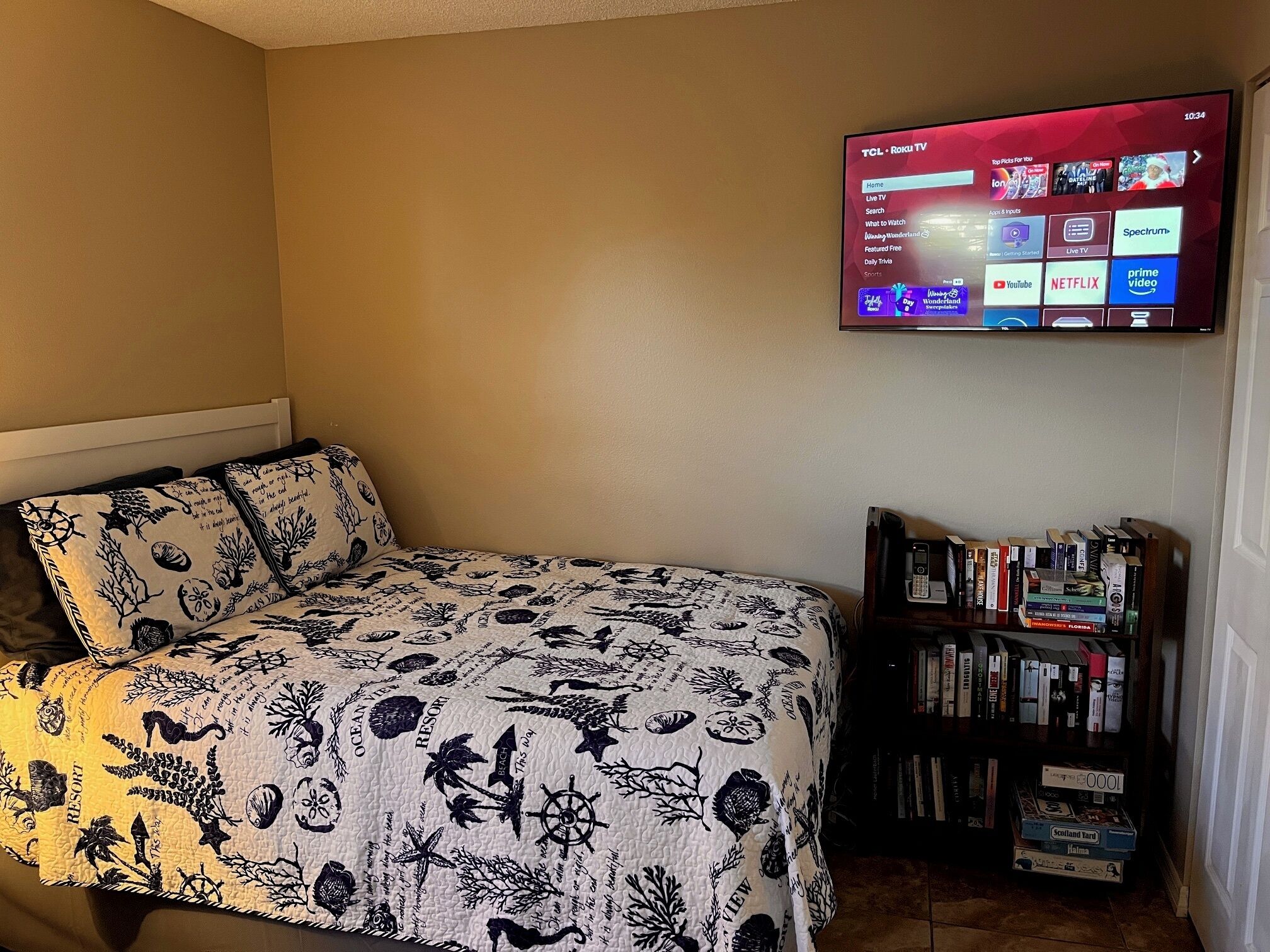 Un dormitori amb un llit, un televisor fixat a la paret que mostra la interfície d'una plataforma de streaming, i una estanteria amb diversos llibres.