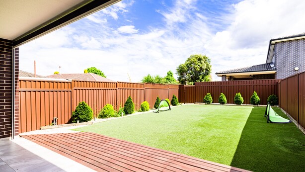 Ein Garten mit Kunstgrasanlage, einer Holzdielenveranda und einem eingezäunten Bereich mit geschnittenen Büschen und einem kleinen Baum.