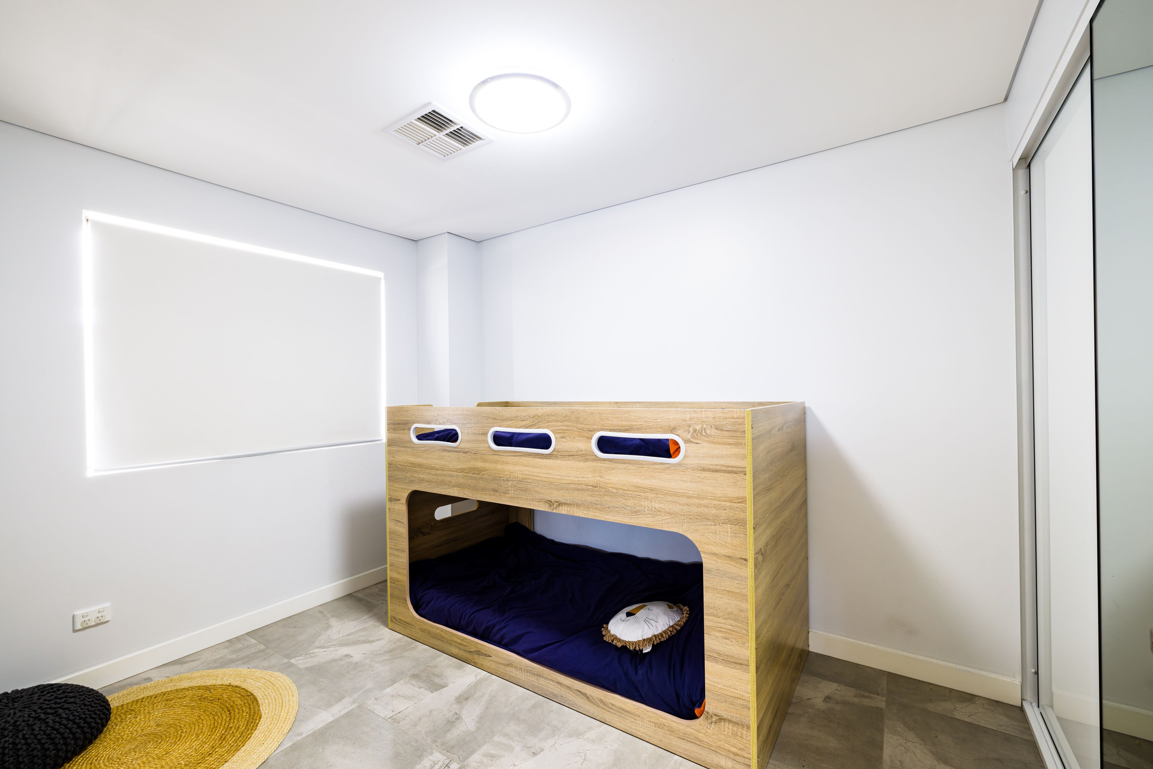 Een stapelbed met een ingebouwd bureau en opbergvakken, een effen witte muur, en een klein vloerkleed.