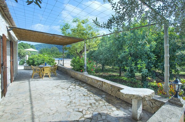 En udendørs terrasse med flisebelægning, et bord med stole og en pergola, der giver skygge.