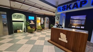 Zona de recepción con piso a cuadros, dos sillones verdes, un mostrador de madera y una pantalla grande que muestra un paisaje.