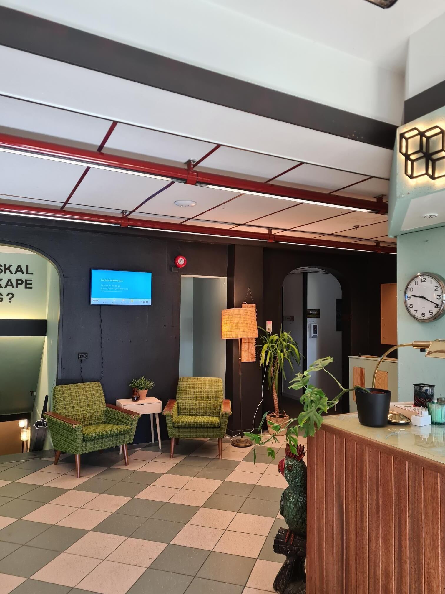 Una moderna zona de recepción con suelo a cuadros, dos sillones verdes, un mostrador de madera, un reloj de pared y una pantalla digital que muestra información.