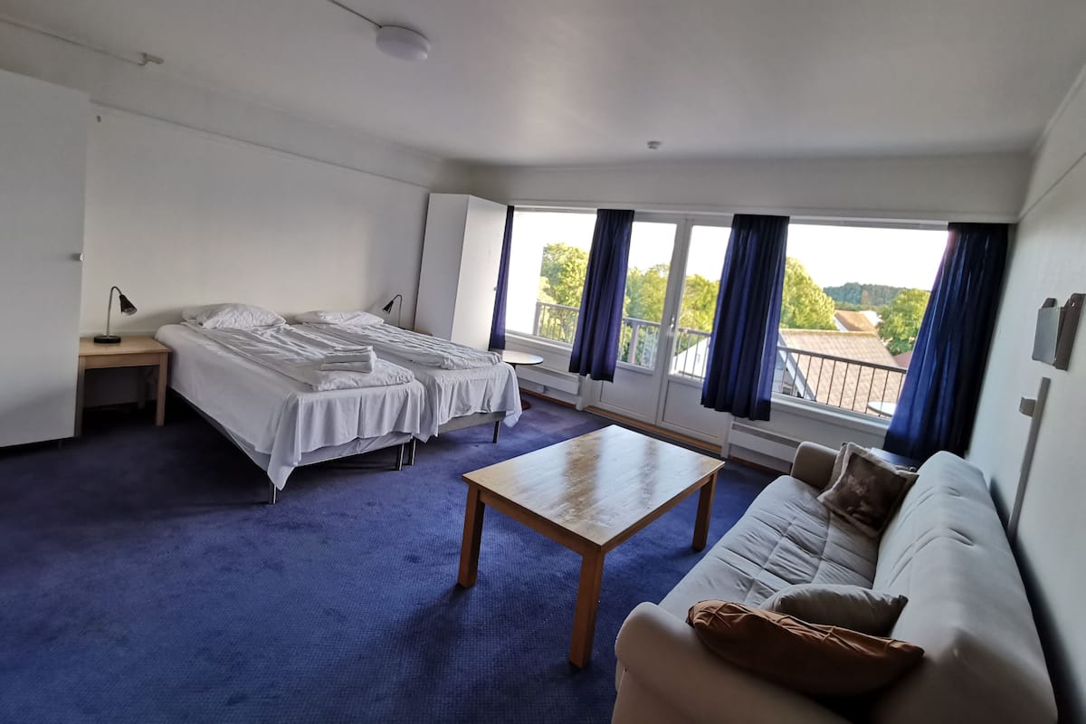 A hotel room with two beds, a sofa, a wooden table, and large windows with curtains.