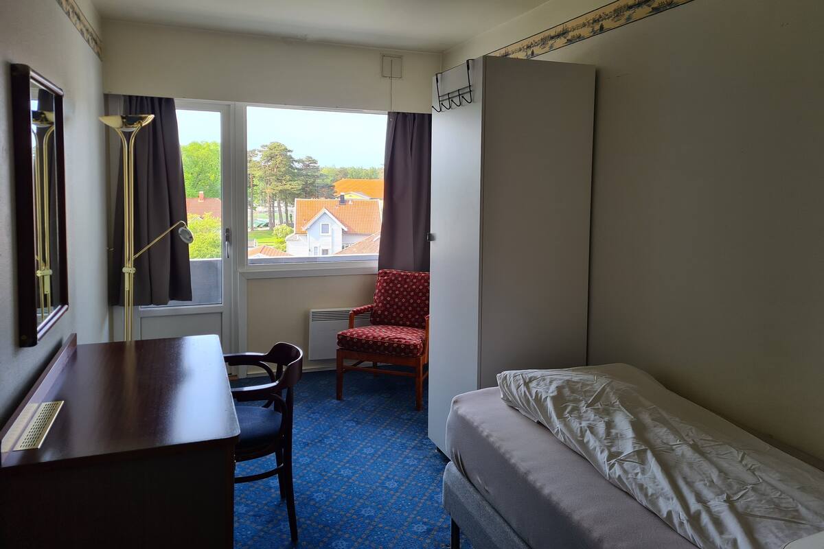 A small, neatly arranged room with a bed, a desk, and a chair. There is a window providing a view of a house and trees.