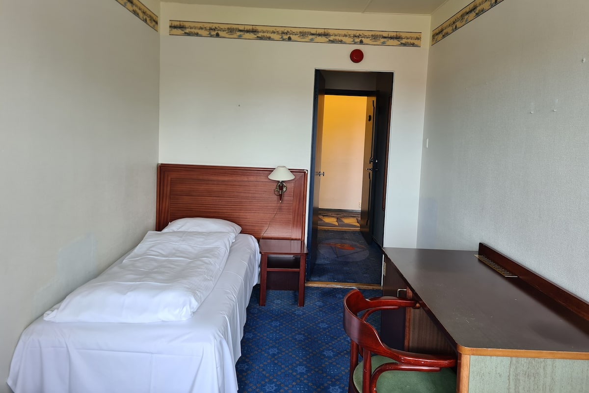 A hotel room with a single bed, a wooden desk, a chair, and a carpeted floor.