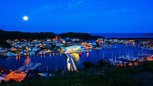 Un pueblo costero de noche con un puerto deportivo, edificios iluminados y cielo despejado.