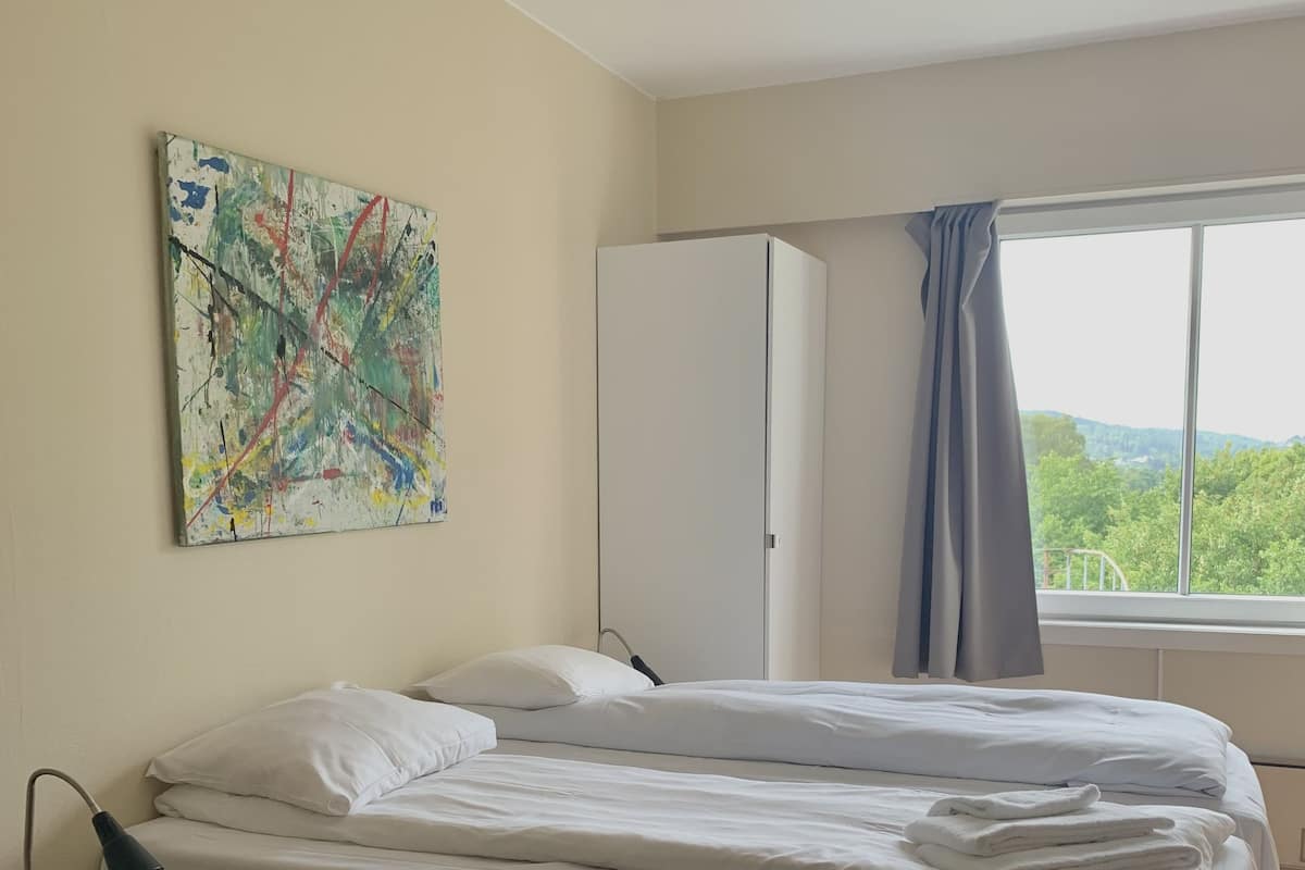 A bedroom with a bed, a nightstand, a desk, a wardrobe, a window with a view of greenery, and a colorful abstract painting on the wall.