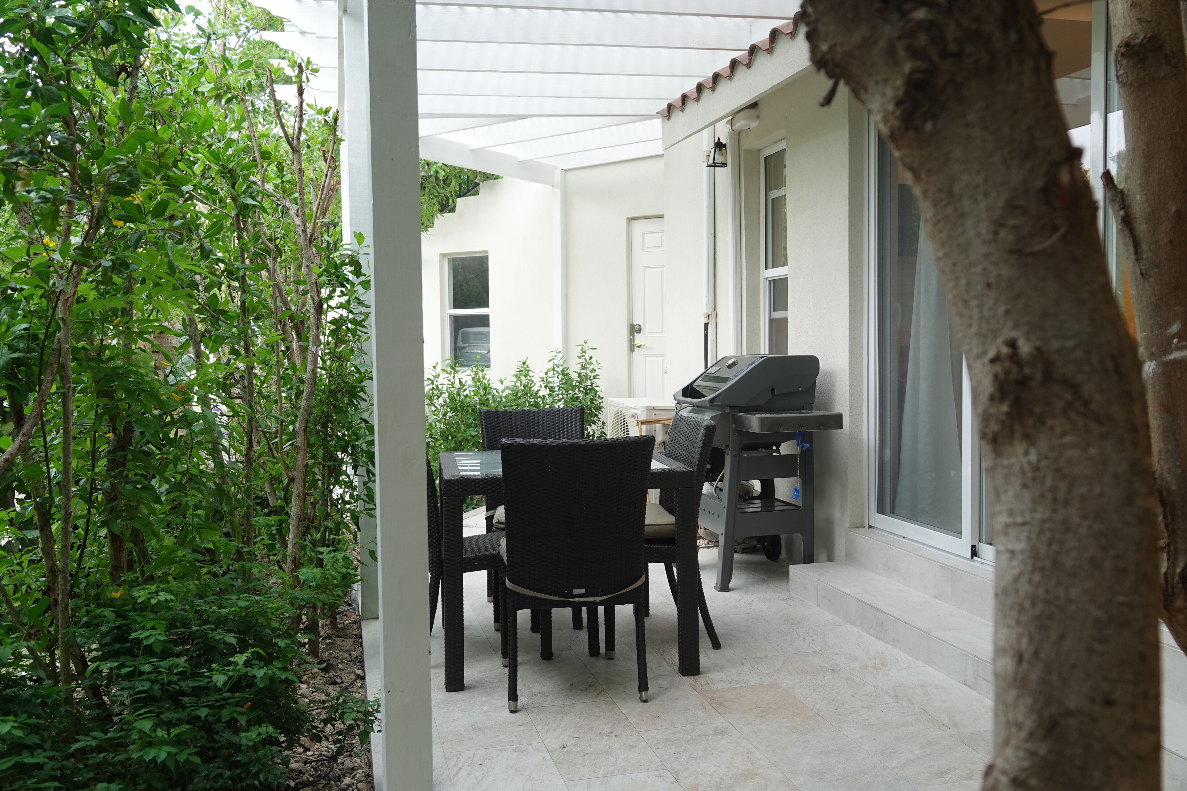 Een terras buiten met een eettafel, stoelen, een barbecue en een witte muur met een deur.