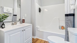 A bathroom with a white sink, mirror, and bathtub. There is a towel rack with a white towel, a potted plant, and toiletries on the counter.