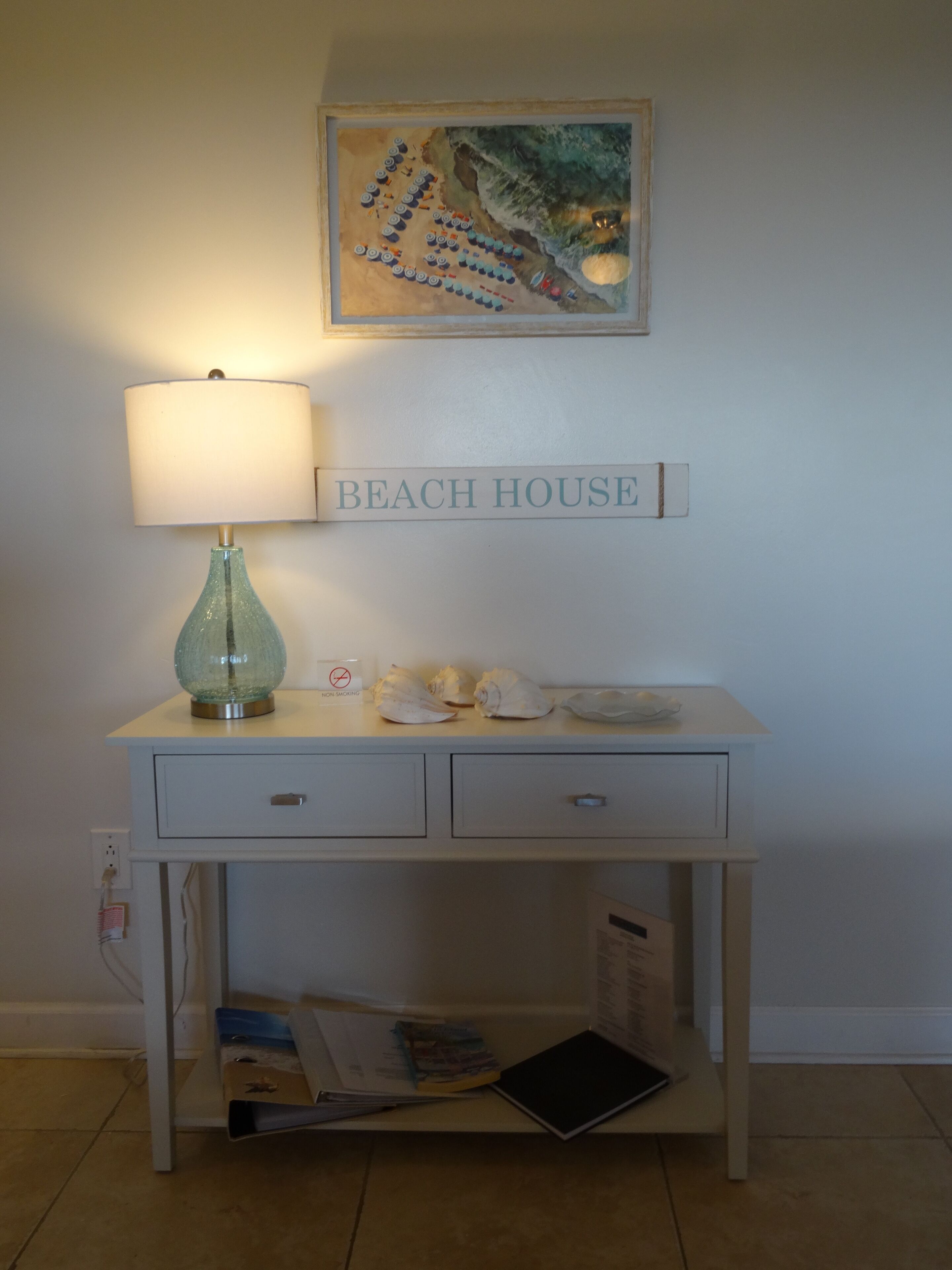A desk with a lamp, a 'BEACH HOUSE' sign, and a painting above.