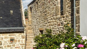 Un bâtiment en pierre avec un toit noir et une fenêtre. On aperçoit un jardin avec des fleurs roses au premier plan.