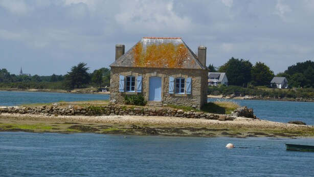 Une maison en pierre avec des volets bleus, située au bord d’une étendue d’eau paisible.