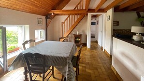 Un coin repas avec une table et des chaises, des poutres en bois au plafond, une vue par la fenêtre et un espace cuisine à l’arrière-plan.